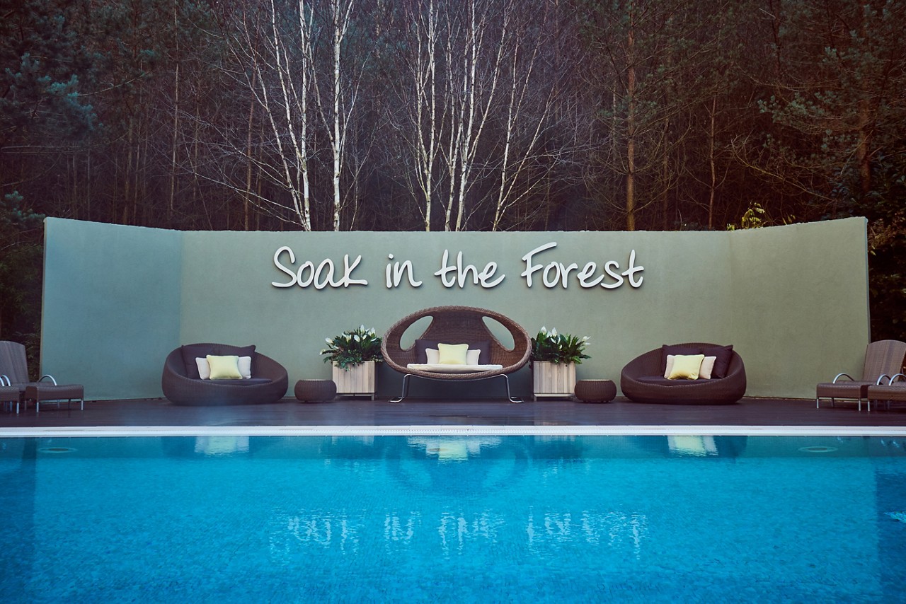 Poolside lounge seating faces a pale wall, displaying the words: “Soak in the Forest,” amid potted plants; wicker chairs and cushions sit on a deck, with a dense forest backdrop.